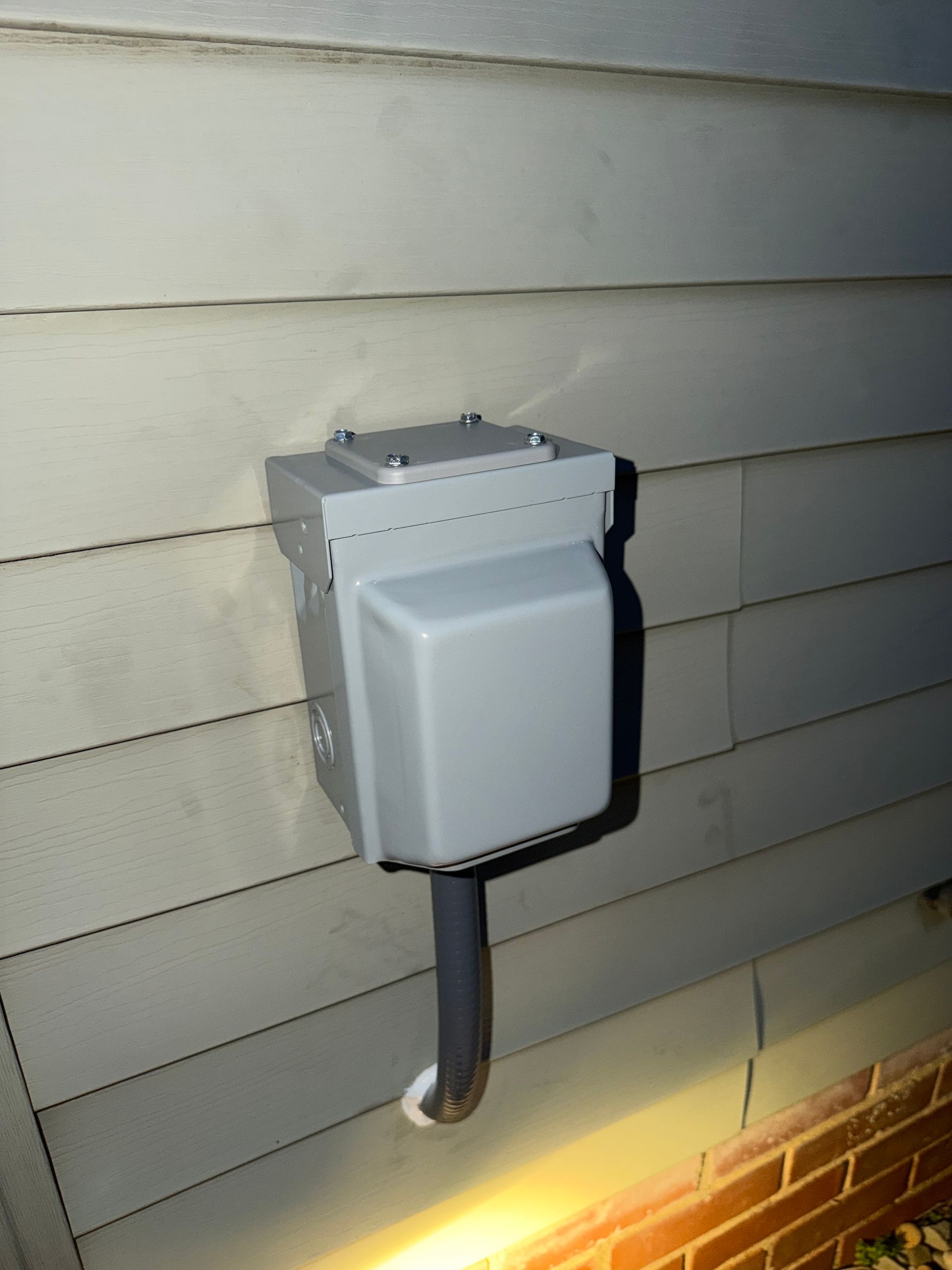 Outdoor electrical box mounted on beige siding above a brick foundation, lit from below.