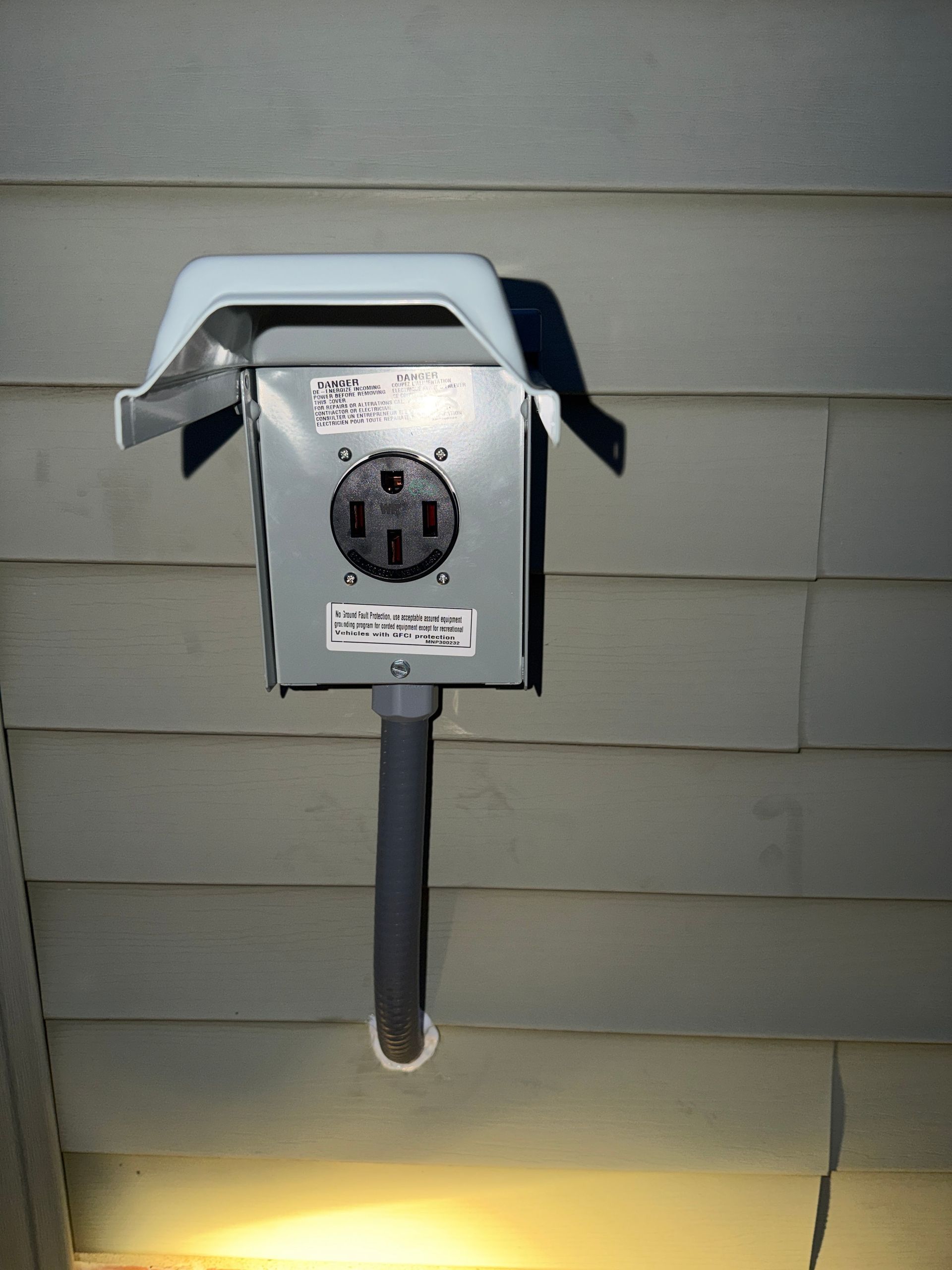 Outdoor electrical outlet box on beige siding with a cover and conduit pipe under a light.