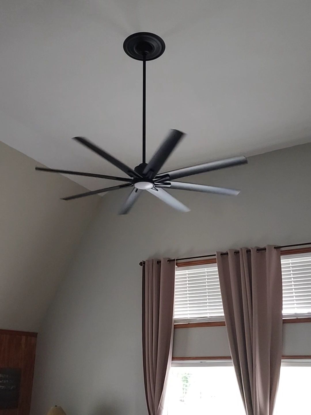 Modern black ceiling fan with long blades hanging in a room above a window with pink curtains.