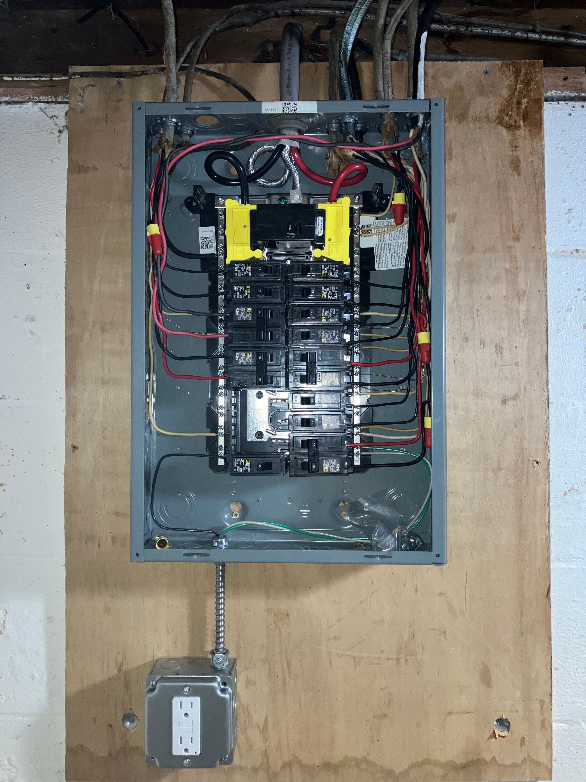 Open electrical breaker panel with exposed wiring and a metal conduit box below on a plywood wall.