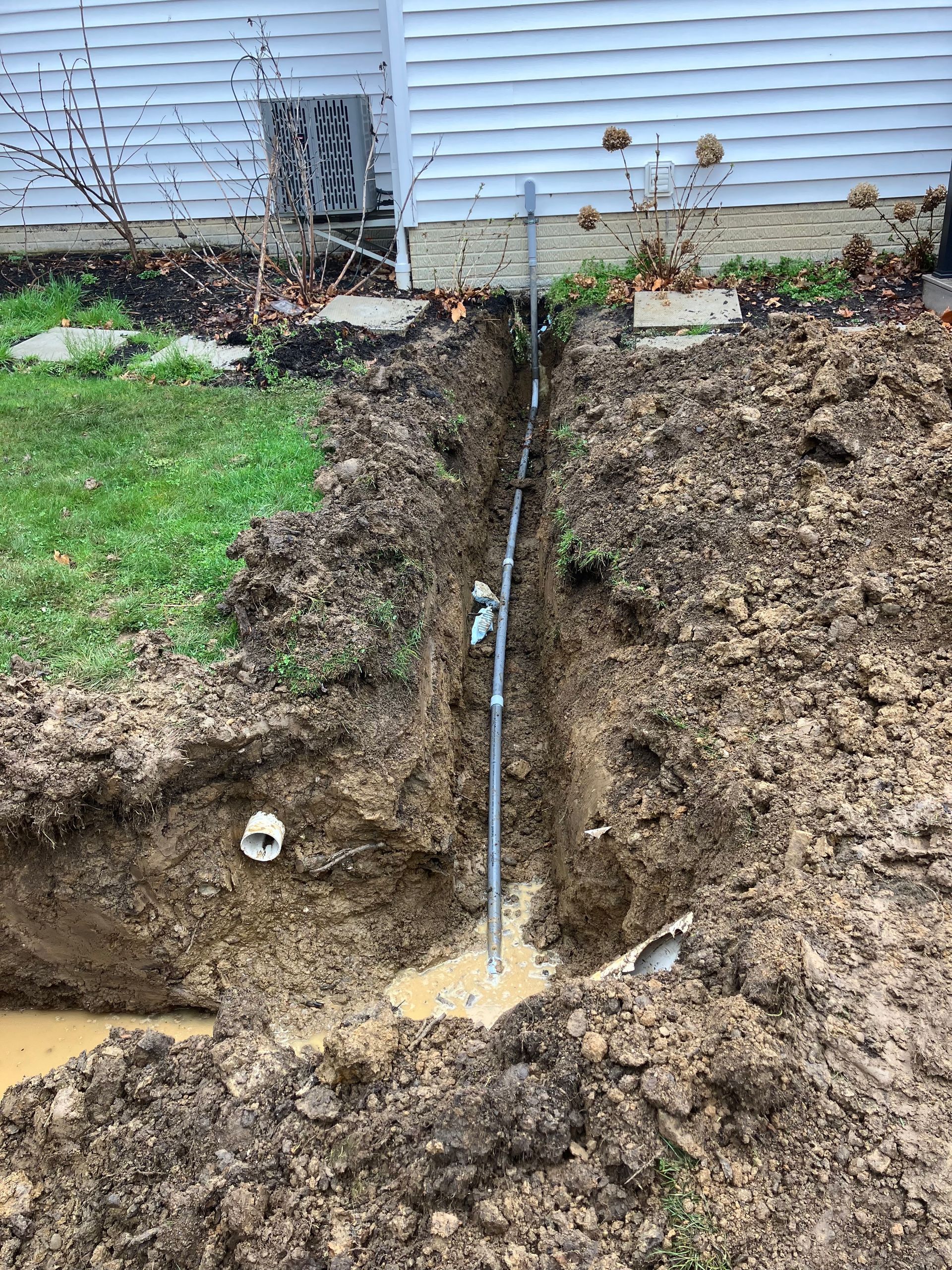 Excavation trench along house foundation exposing a pipe and muddy soil beside patchy grass
