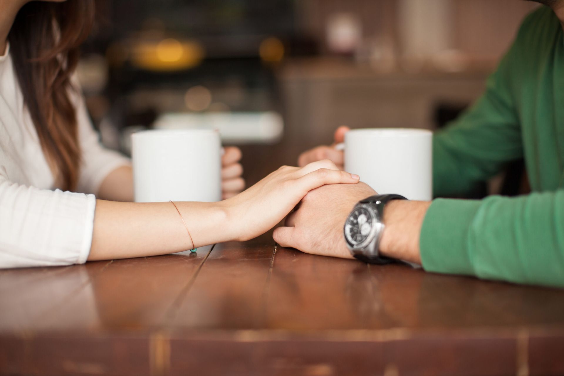 Online Dating | Two people holding hands across a wooden table with coffee mugs in the background