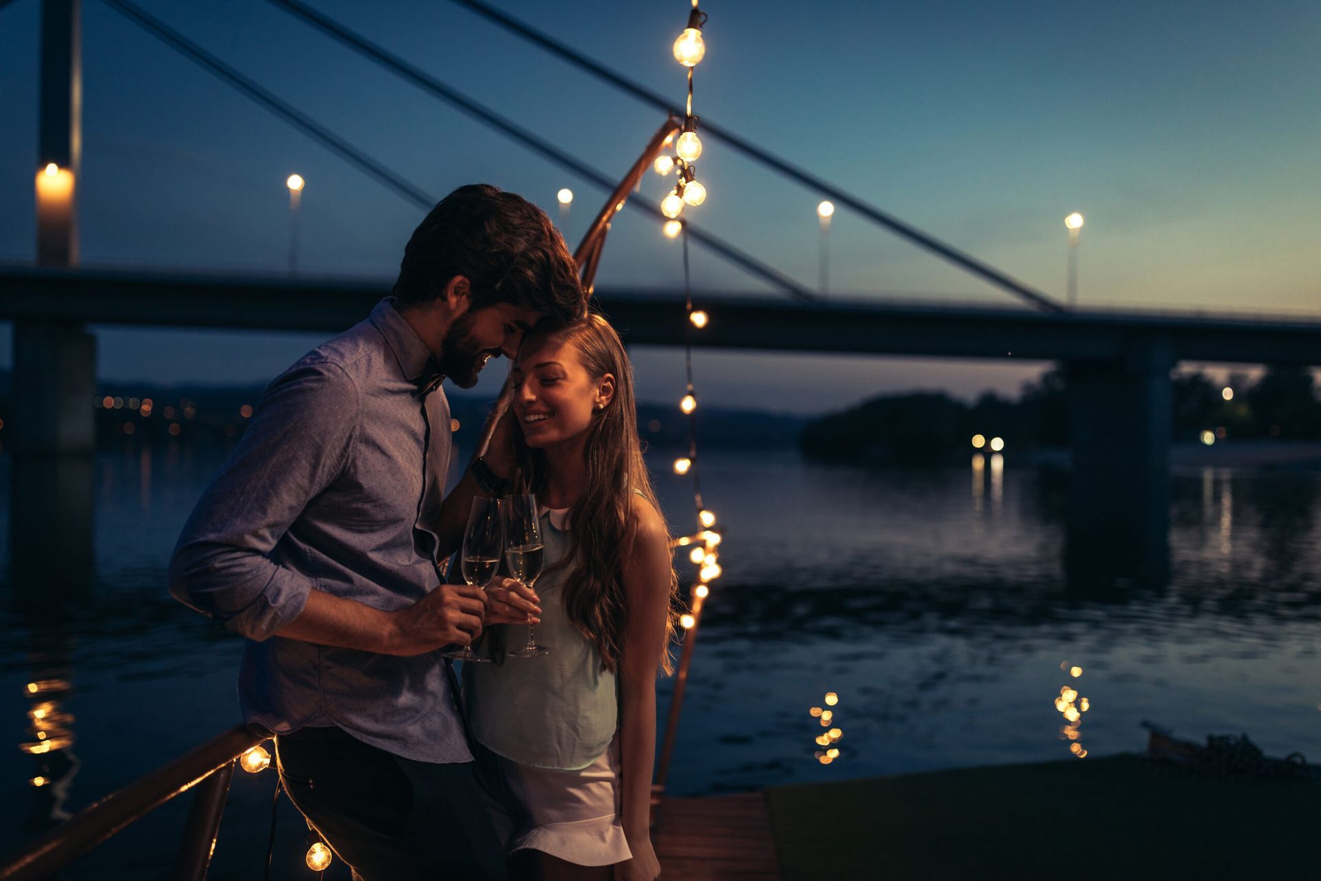 Couple embracing by a river at dusk under string lights, with a bridge in the background
