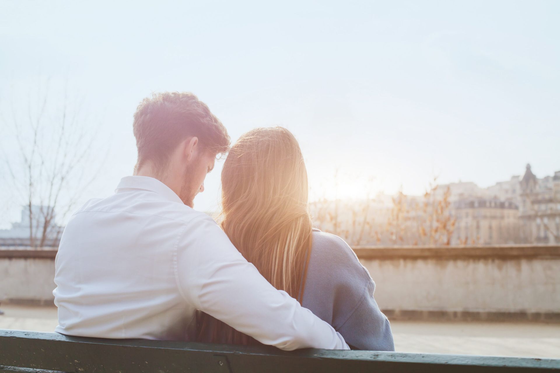 Couple sitting on a bench by the water at sunset, with city buildings in the distance.