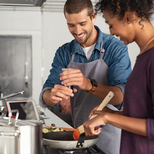 Two people cooking together in a kitchen, stirring a pan and smiling.