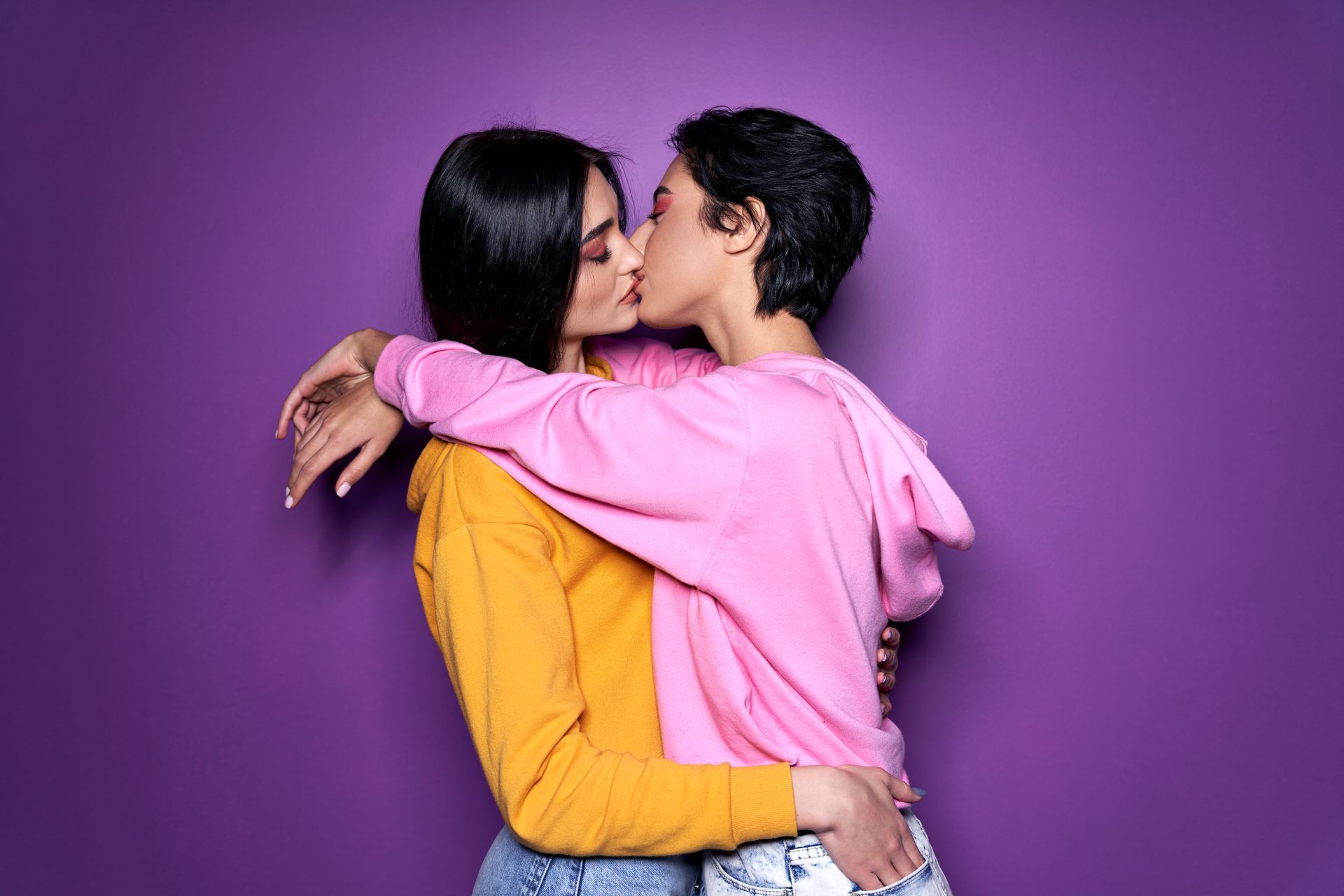 Two people kissing and hugging against a purple background, one in pink and one in yellow.