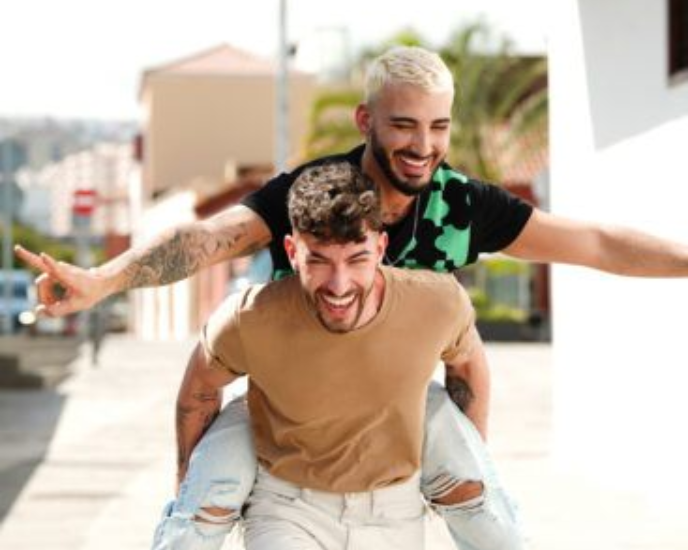 Two smiling friends, one giving the other a piggyback ride outdoors with arms outstretched.