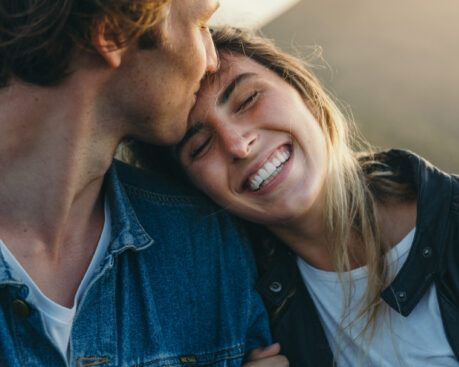 Two people smiling and cuddling outdoors, one kissing the other’s forehead.