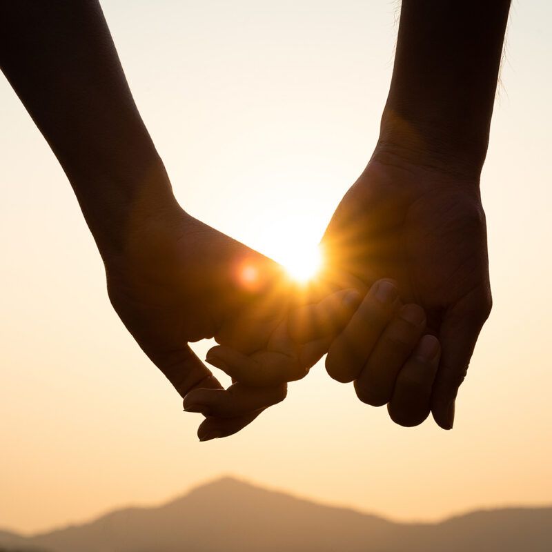 Two hands holding at sunset, with the sun glowing between fingers over distant hills