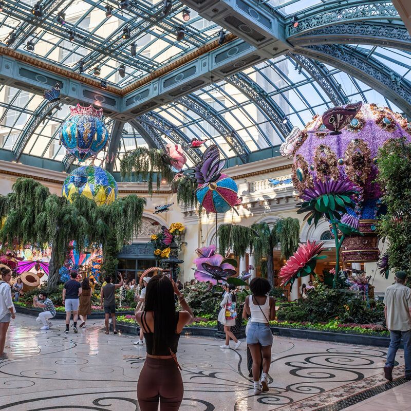 Indoor garden atrium with colorful hanging floral sculptures and visitors walking beneath a glass roof