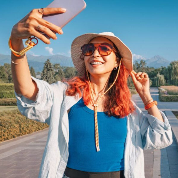 Woman taking a selfie on a sunny road, wearing a hat and sunglasses with mountains in the background