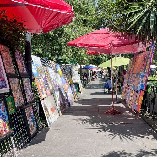 Outdoor art market with colorful paintings displayed under red umbrellas along a tree-lined walkway