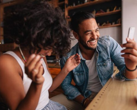 Two people smiling and laughing while looking at a phone at a wooden table indoors