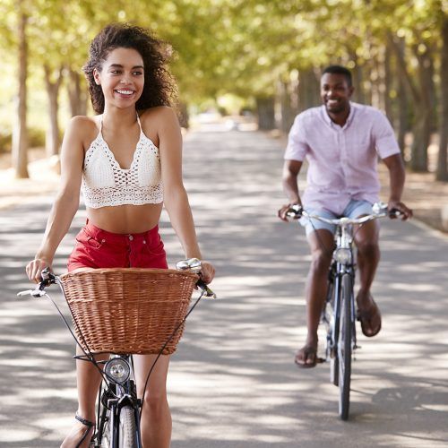 Two people riding bicycles on a sunny tree-lined path, one smiling in front with a wicker basket