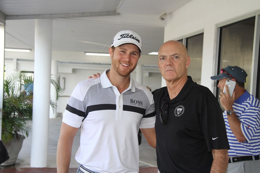 Two men, one with a golf hat and polo, the other with a bald head and black shirt, pose for a photo.