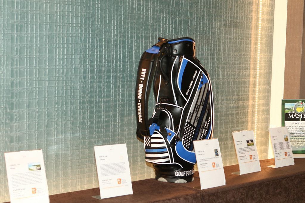 Black, blue, and white golf bag displayed on a shelf in front of a textured wall with price tags.