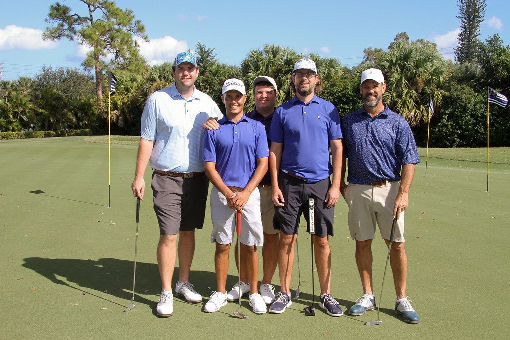 Five men in golf attire stand on a green, smiling at the camera. Sunny day.