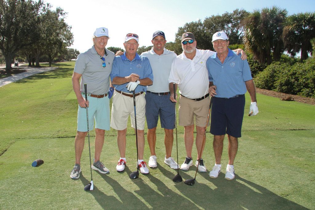 Five men in golf attire pose on a golf course. They are smiling, holding clubs, and in sunny conditions.