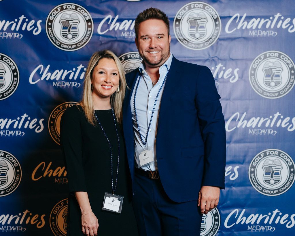 A man and woman pose in front of a blue backdrop. The man wears a blue suit, the woman, a black dress. Both smile.