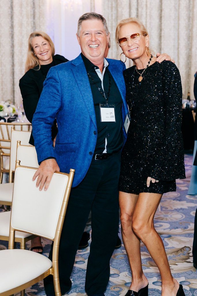 Man in blue jacket smiles, hugs woman in black dress. Woman looks at the camera, and another woman stands behind.