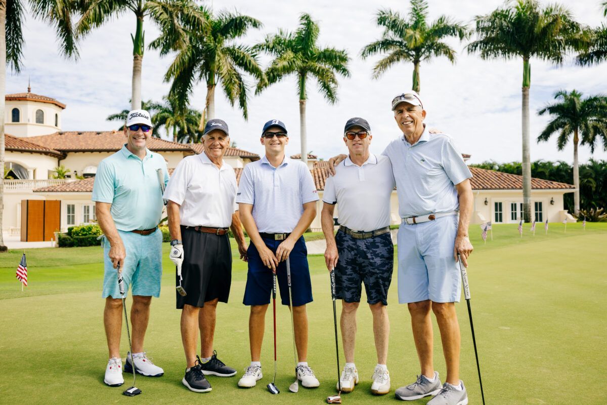 Five men in golf attire pose on a green. Palm trees and a large building are in the background.