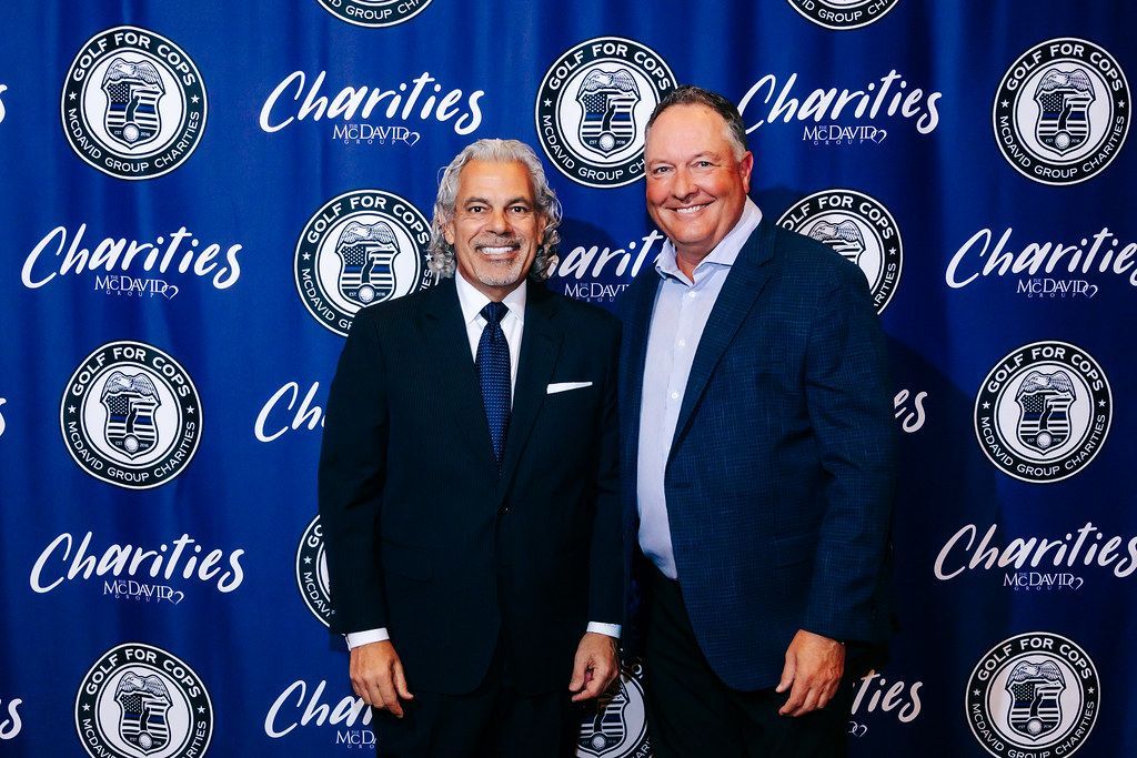 Two men smiling, posed in front of a blue backdrop with a logo that reads