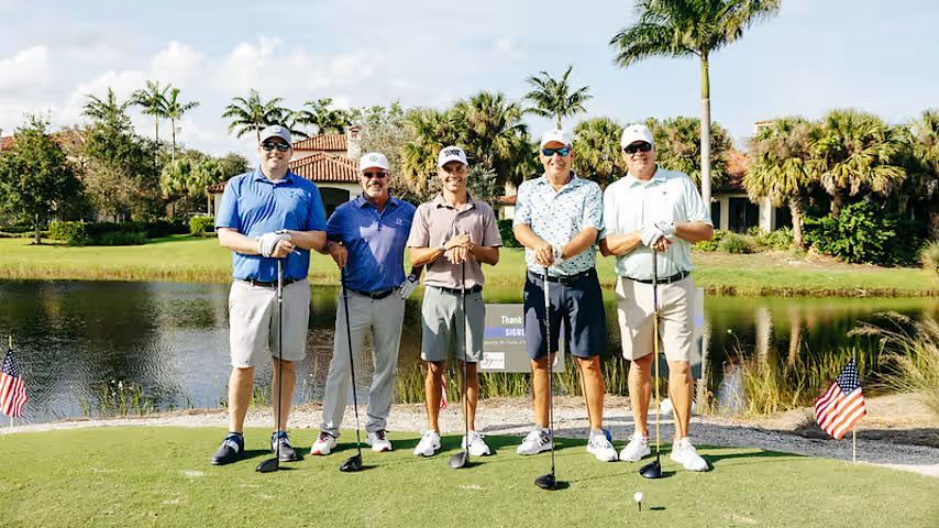 Five men stand on a golf course, smiling with golf clubs. They are by a lake on a sunny day.
