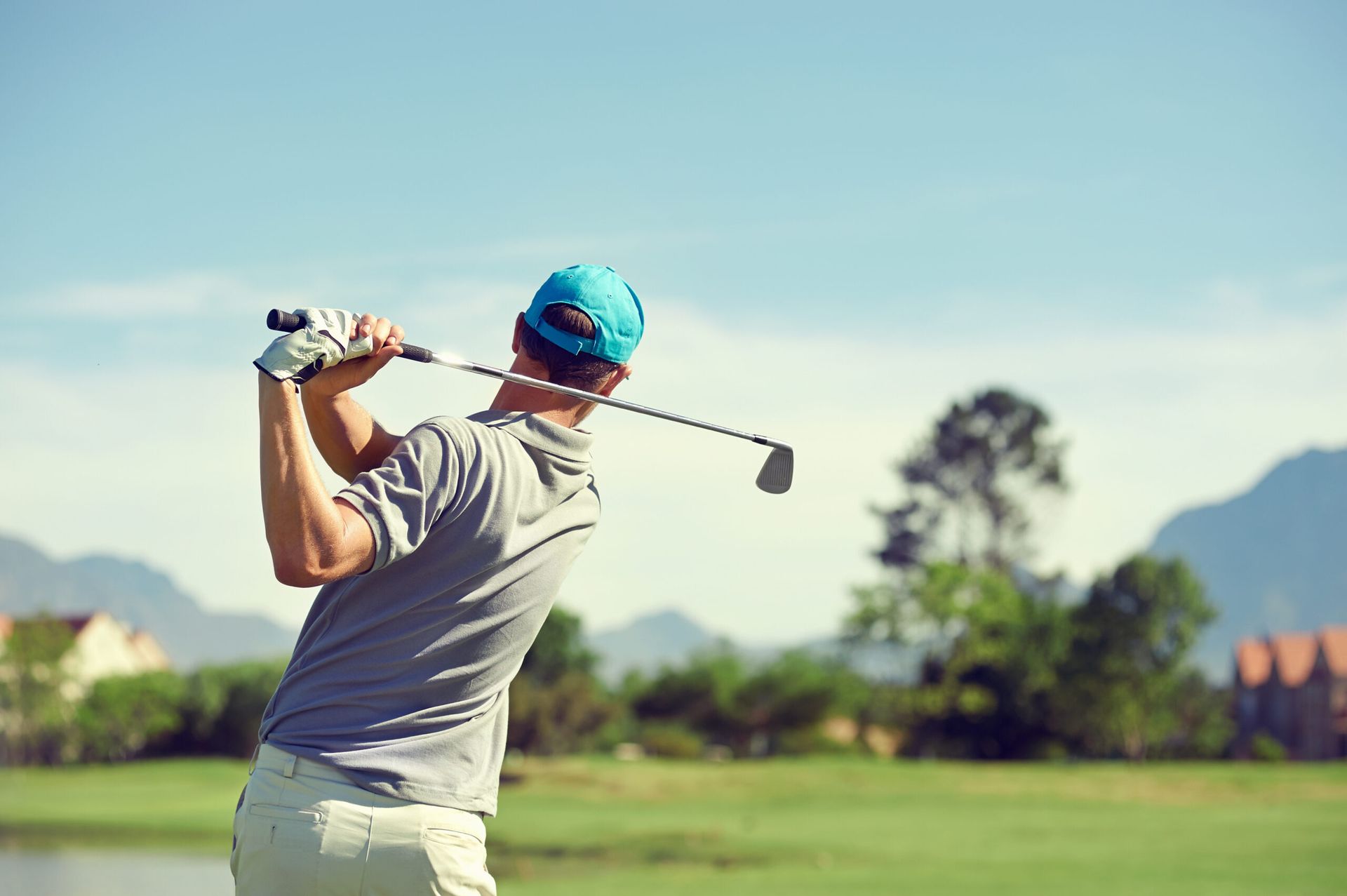 Golfer in gray shirt and blue hat swings club on green course with mountains in the background.