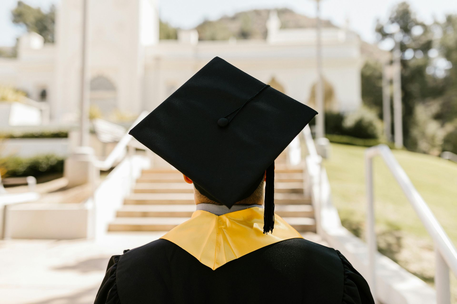 Graduation: Person in cap and gown walks towards a building with stairs, facing away.