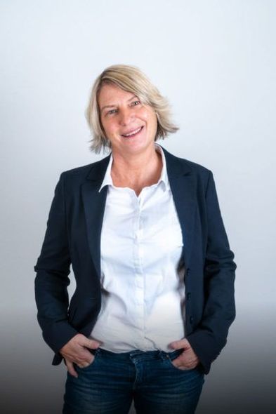 Eine Frau in einer blauen Jacke und einem weißen Hemd steht mit den Händen in den Taschen da.