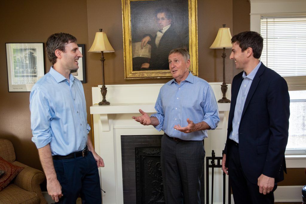 Three men in shirts stand in a room, with the center man gesturing while speaking, in front of a fireplace and portrait.