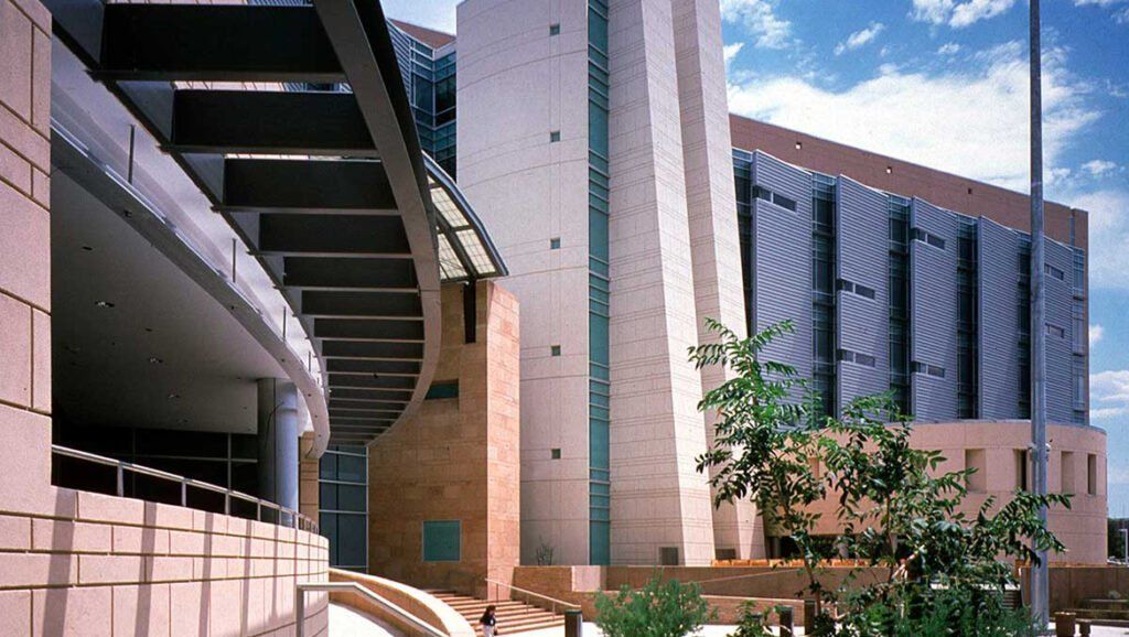 Modern stone and glass building with a curved overhang, metallic paneling, and landscaping under a blue sky.