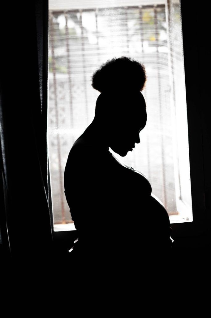 A silhouette of a pregnant person standing in front of a bright window, looking down at their belly.