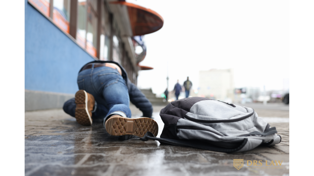 A person in jeans and brown shoes has fallen on a wet, paved walkway near a building, with their backpack lying nearby.