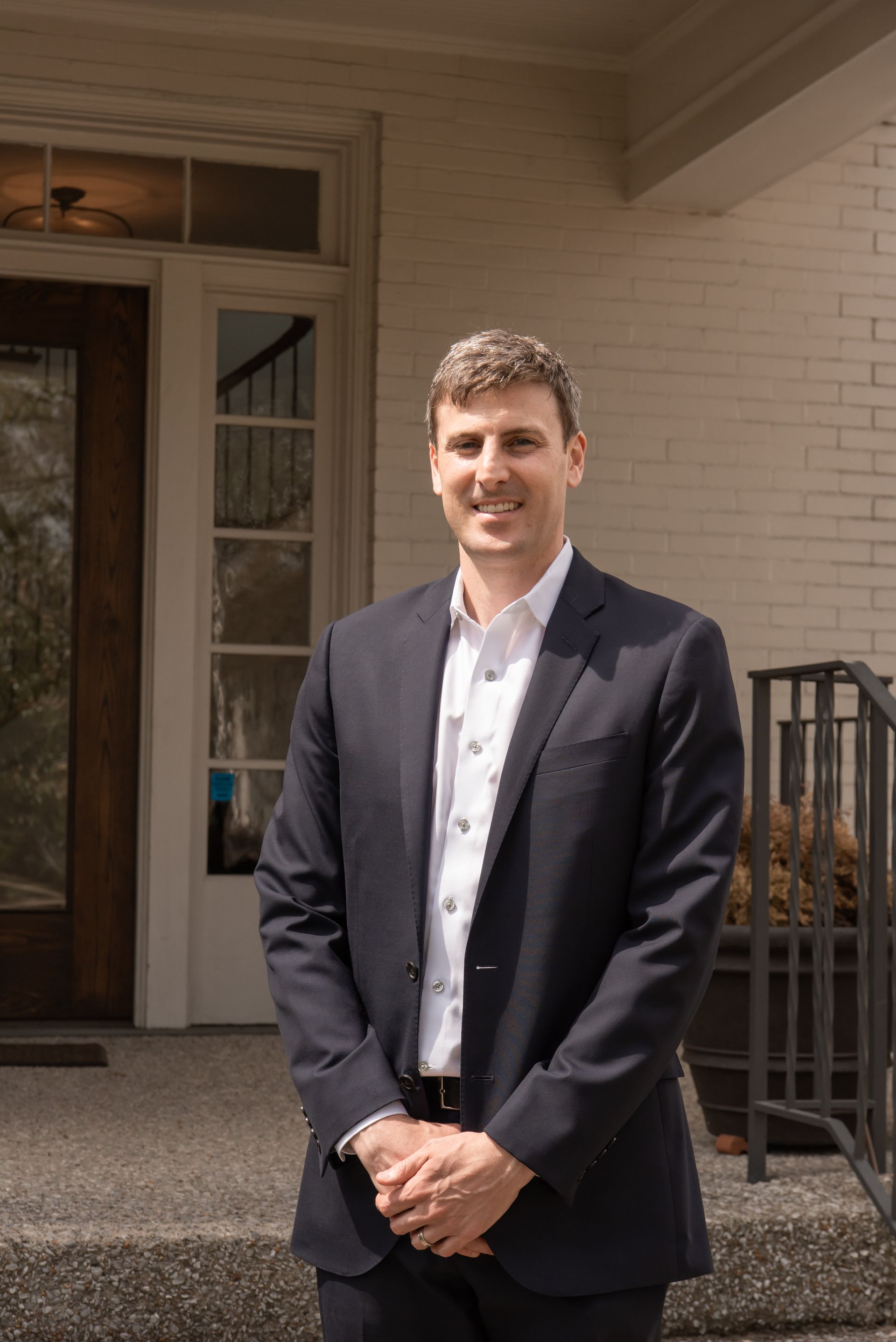 A person wearing a suit and white shirt standing outdoors in front of a building with a glass door and brick exterior.