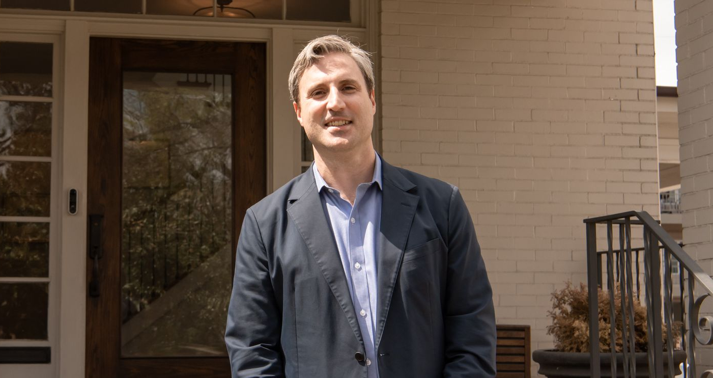 Man in a blue suit smiles, seated in front of a fireplace.