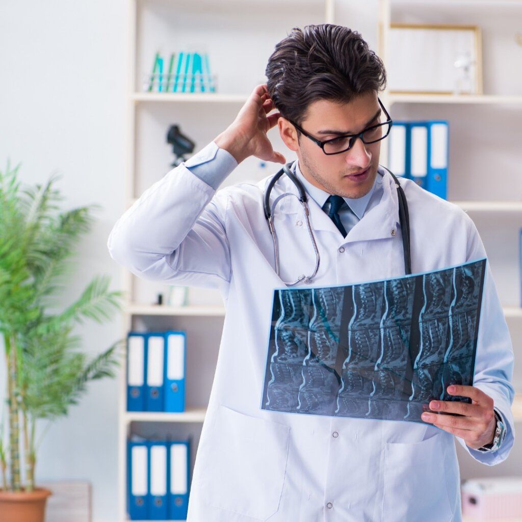 A doctor in a white coat with a stethoscope around their neck scratches their head while examining an X-ray.
