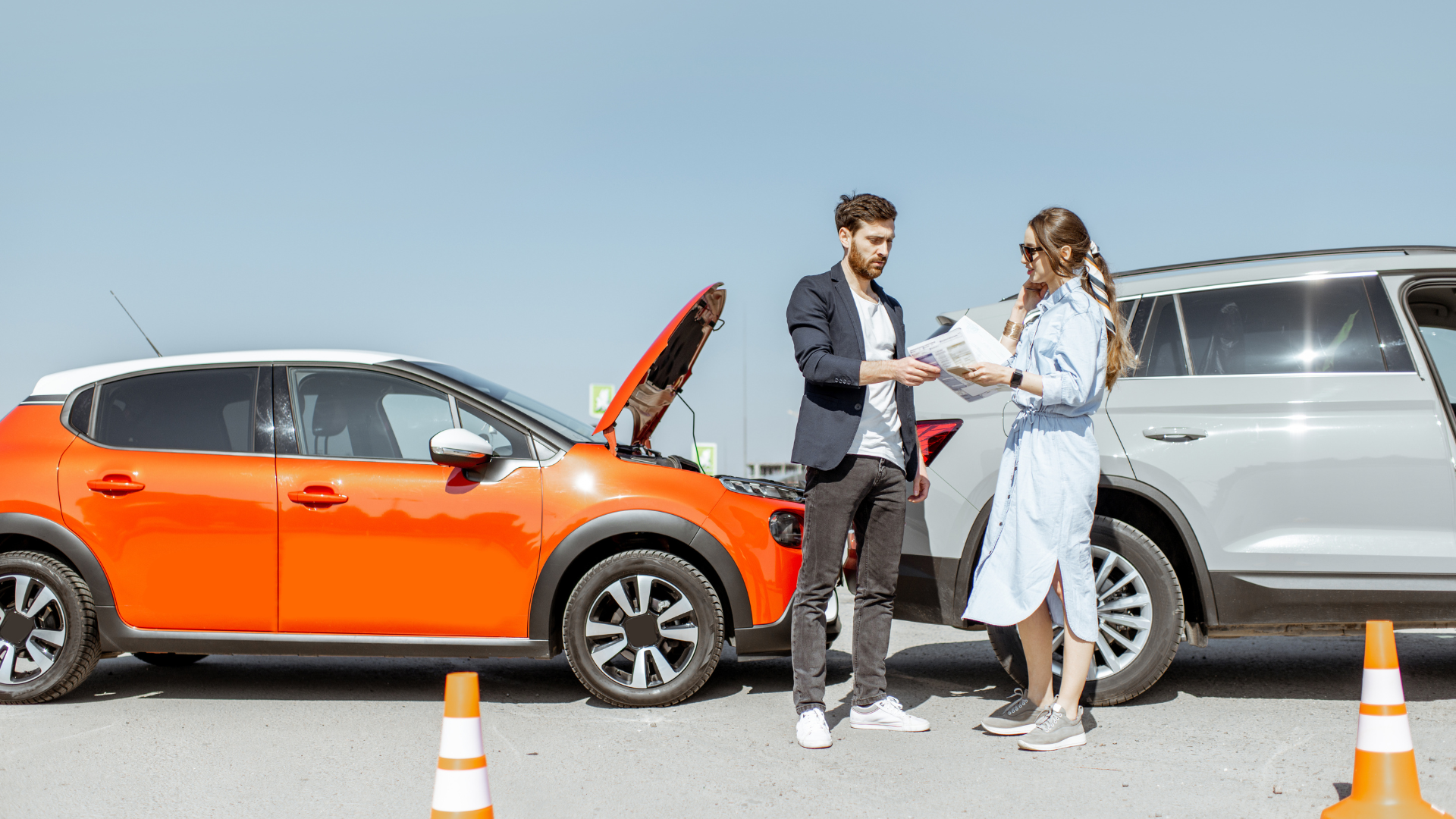 Two people stand between an orange car with its hood up and a grey SUV, exchanging insurance documents outdoors.