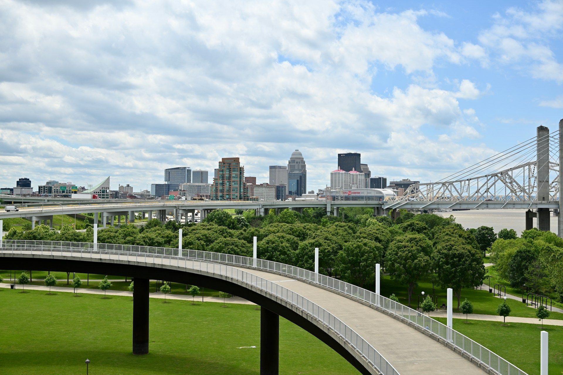 A curved pedestrian bridge leads toward a lush green park with a modern city skyline and a large steel bridge in back.