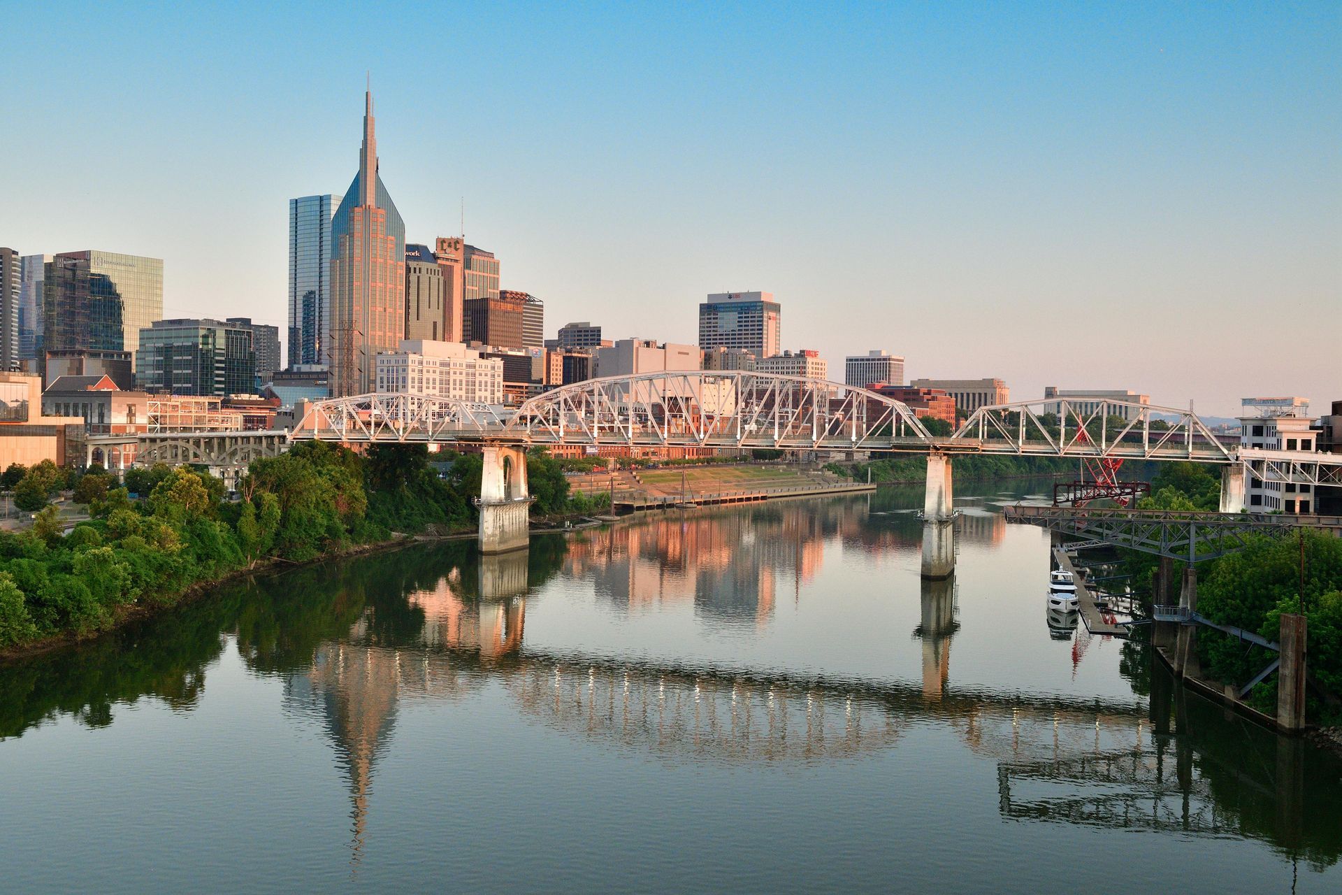 A scenic view of the Nashville skyline and the historic Shelby Street Bridge reflected in the Cumberland River at sunset.