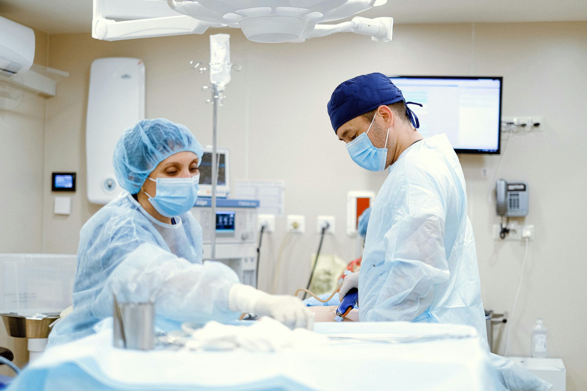 Two medical professionals in surgical scrubs, caps, and face masks work at a table in a bright, sterile operating room.