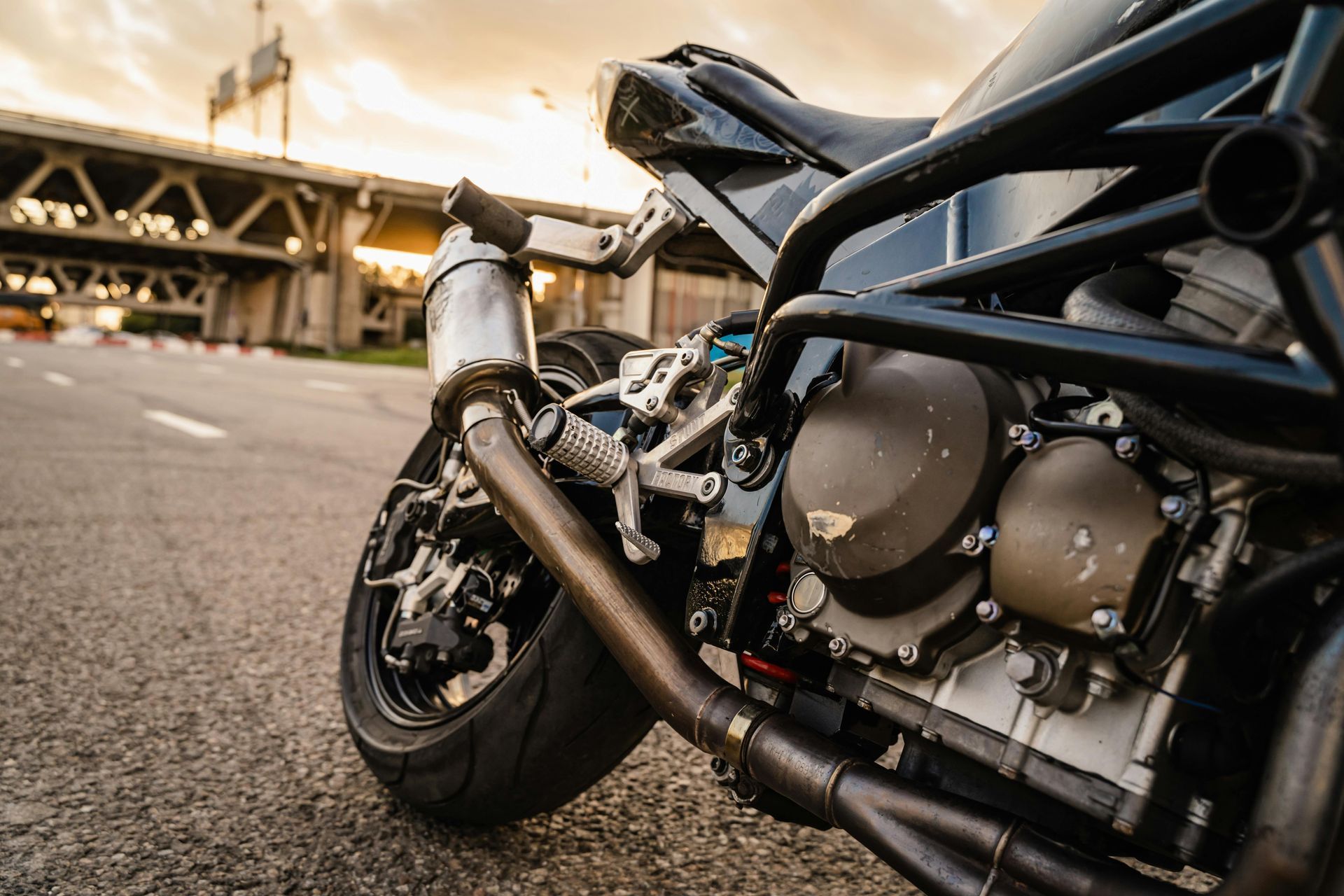 Close-up of a dark-colored motorcycle parked on an asphalt road with an overpass visible in the background at sunset.