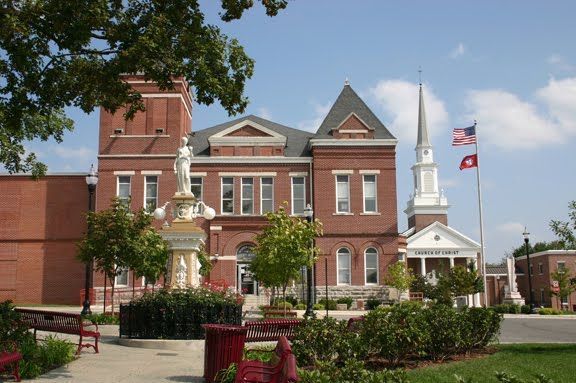 Red brick courthouse with a central statue, side tower, and a church steeple in the background on a sunny day.