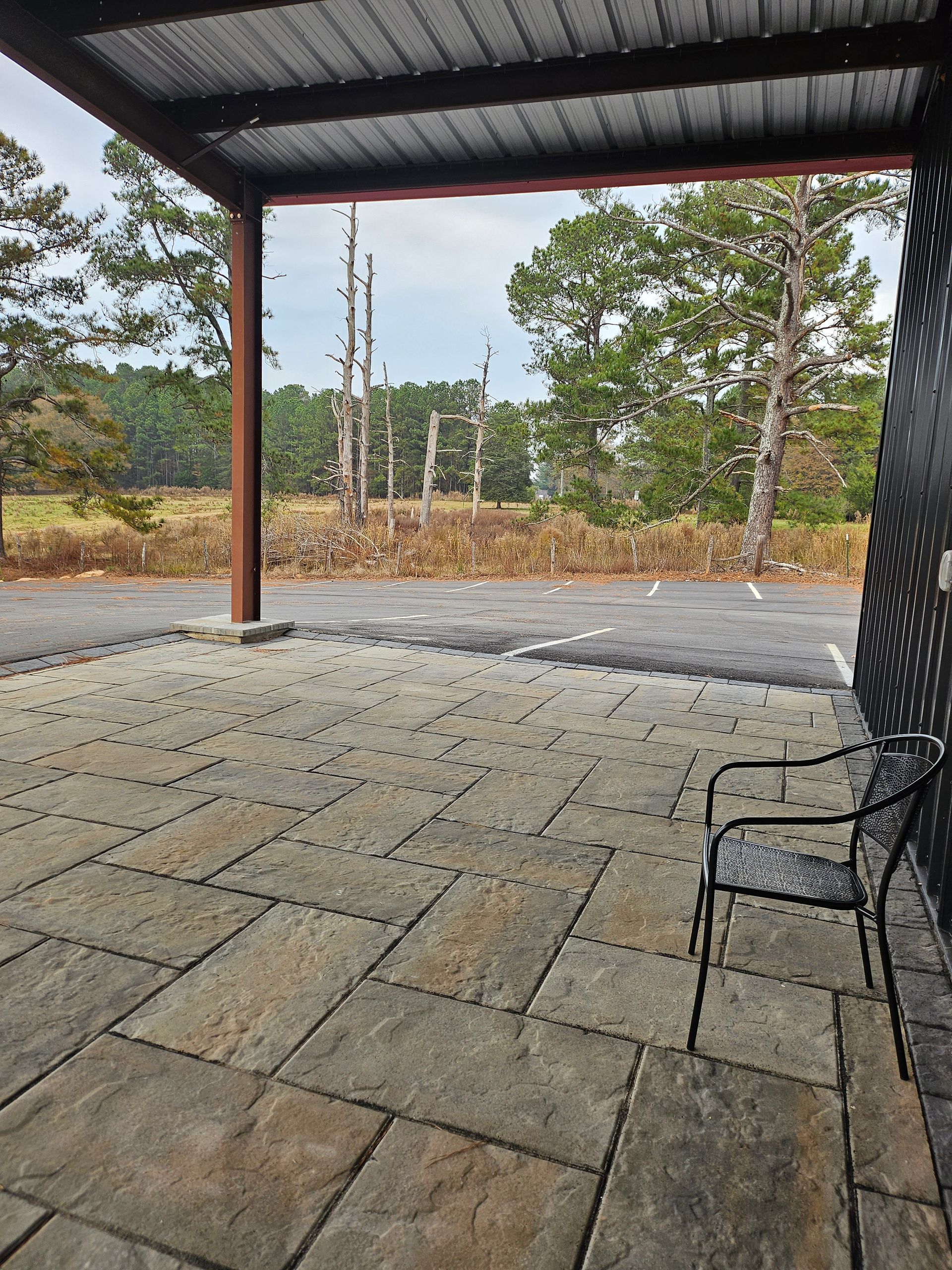 Covered patio with stone floor, black metal chair, and view of trees and gravel outside.