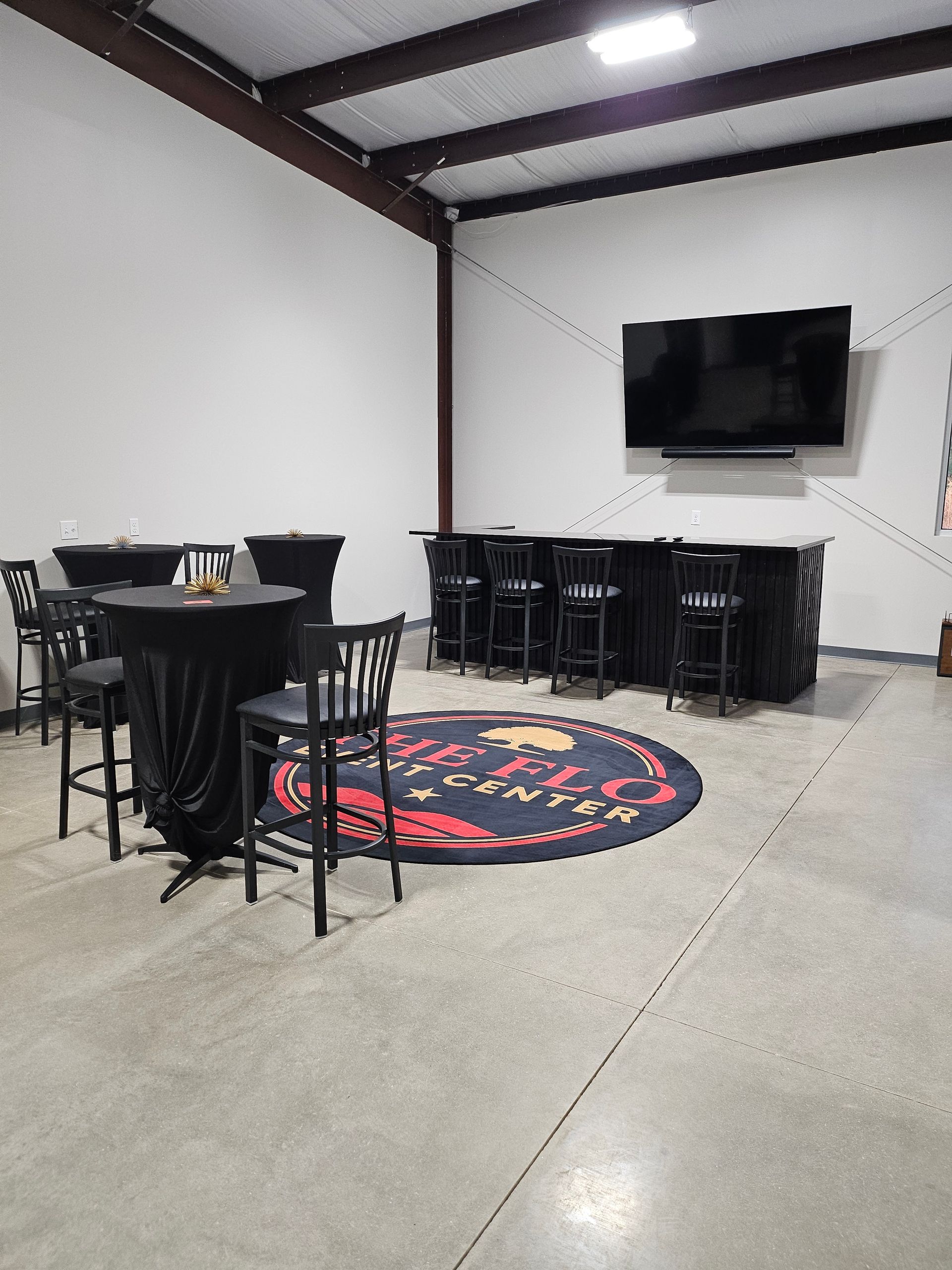 Indoor event room with round tables, black chairs, a wall-mounted TV, and a decorative rug.