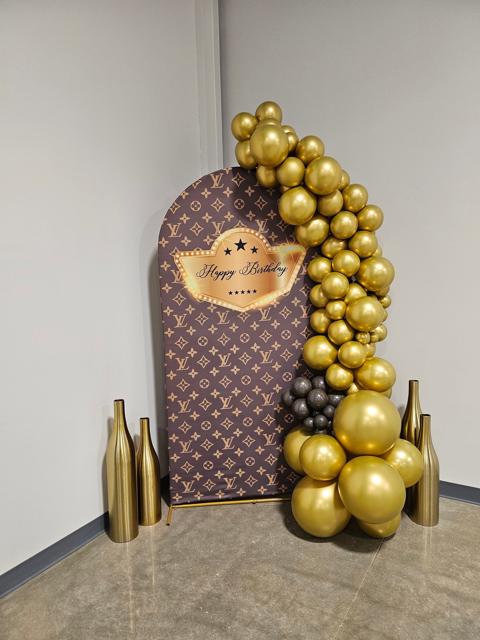 Gold balloon arch beside patterned backdrop with gold bottles on carpeted floor
