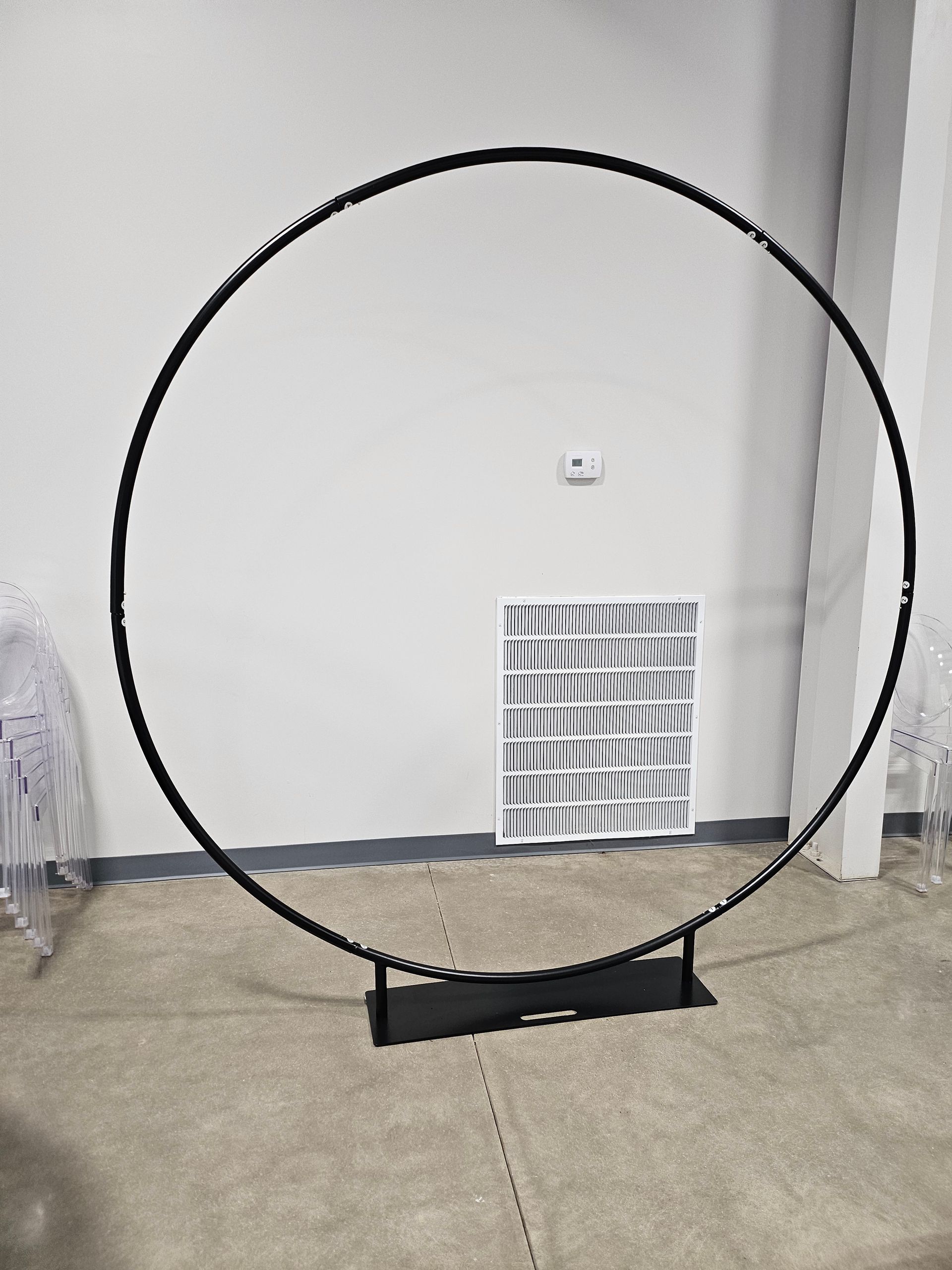Black circular frame standing on a base in front of a white wall and vent on carpet.