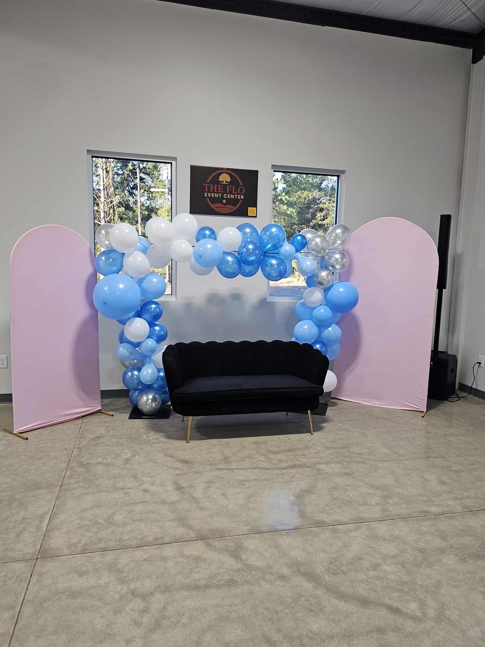 Pastel blue and pink balloon arch framing a black loveseat in an empty event space