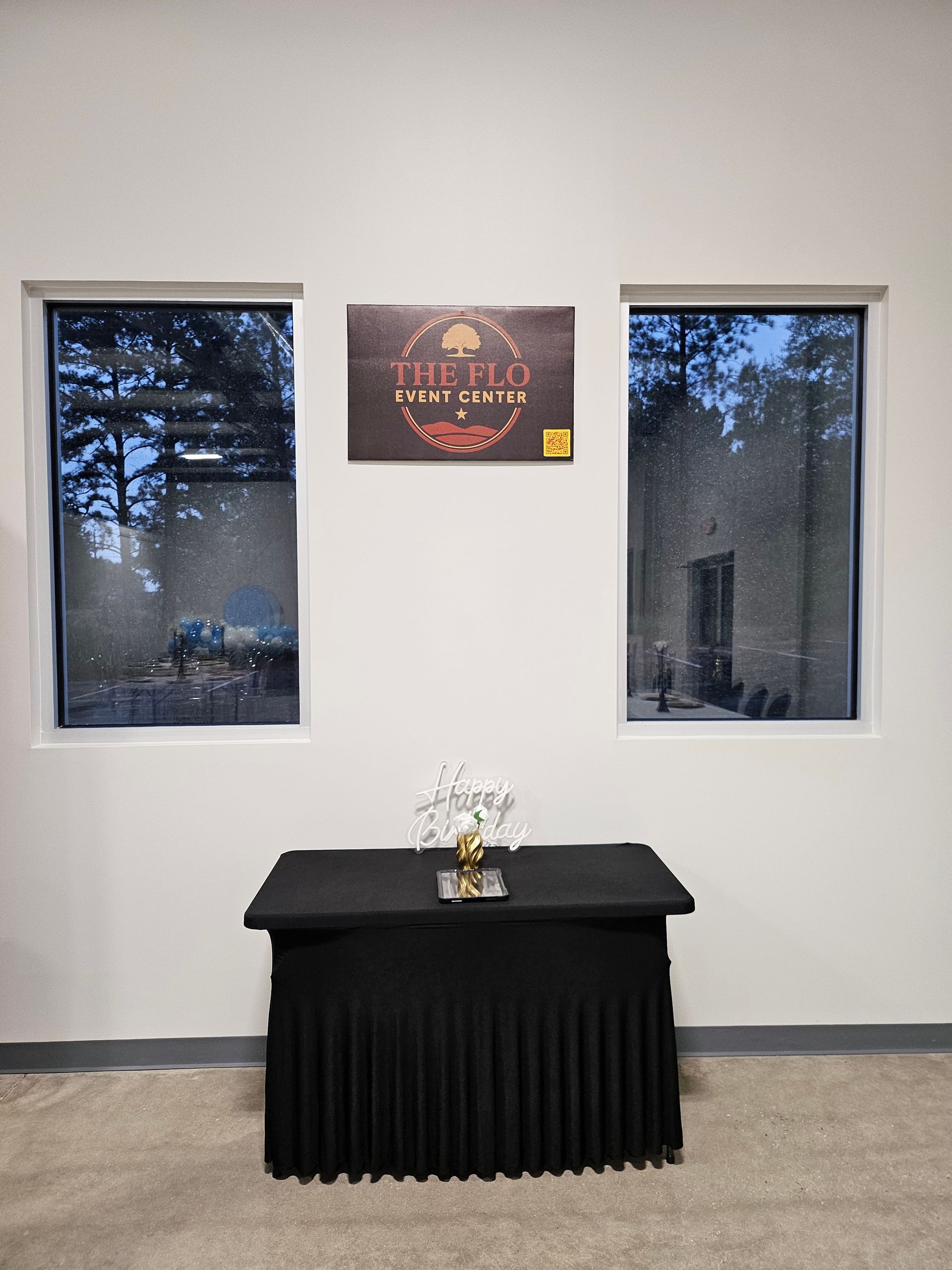 Small memorial table with flowers beneath two windows and a wall sign in a simple room