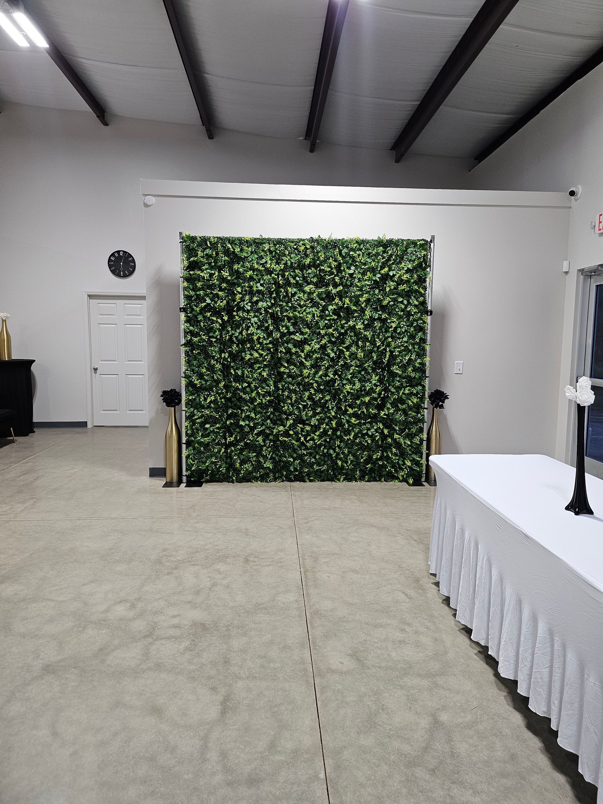 Indoor event space with a green leafy backdrop, white table, and open floor area